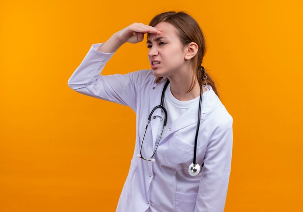 Недовольная молодая женщина-врач в медицинском халате и стетоскопе, положив руку ко лбу и глядя вдаль на изолированной оранжевой стене