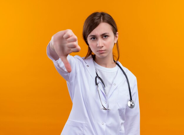 Unpleased young female doctor wearing medical robe and stethoscope showing thumb down on isolated orange wall with copy space