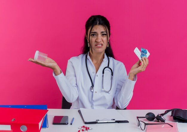 Unpleased young female doctor wearing medical robe and stethoscope sitting at desk with medical tools holding medical drugs and beaker  isolated on pink wall