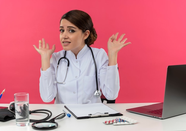 Unpleased young female doctor wearing medical robe and stethoscope sitting at desk with medical tools and laptop showing empty hands isolated on pink wall