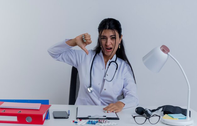 Foto gratuita giovane medico femminile dispiaciuto che indossa veste medica e stetoscopio seduto alla scrivania con strumenti medici mettendo la mano sulla scrivania che mostra il pollice verso il basso cercando isolato