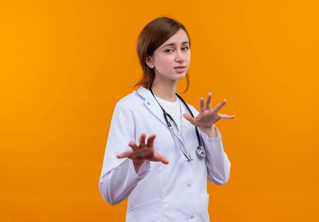 Unpleased young female doctor wearing medical robe and stethoscope stretching hands  on isolated orange wall with copy space