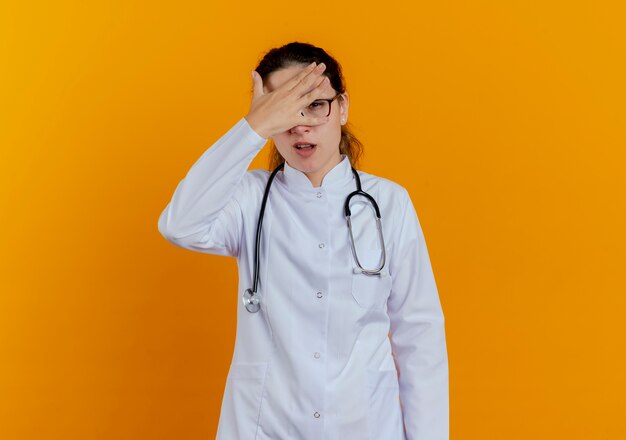 Недовольная молодая женщина-врач в медицинском халате и стетоскопе в очках, положив руку на изолированный лоб