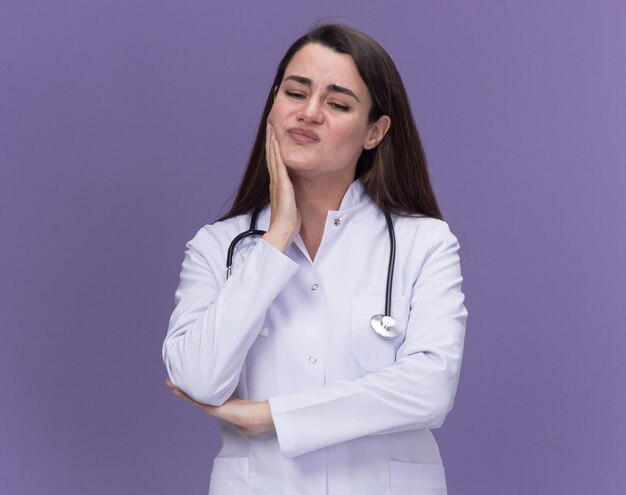 Unpleased young female doctor wearing medical robe with stethoscope puts hand on chin and looks down isolated on purple wall with copy space