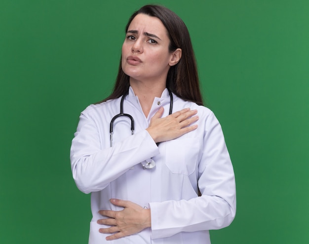 聴診器で医療ローブを身に着けている不機嫌な若い女性医師は、コピースペースで緑の壁に隔離された肩に手を置きます