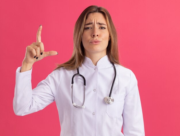 Unpleased young female doctor wearing medical robe with stethoscope showing size isolated on pink wall