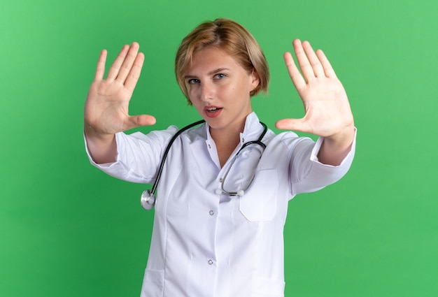 Giovane dottoressa scontenta che indossa un abito medico con uno stetoscopio che mostra il gesto di arresto isolato su sfondo verde