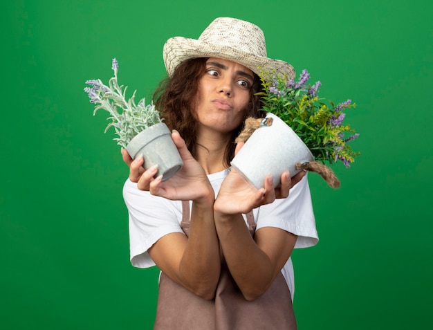 Free photo unpleased young female gardener in uniform wearing gardening hat holding and looking at flowers in flowerpots