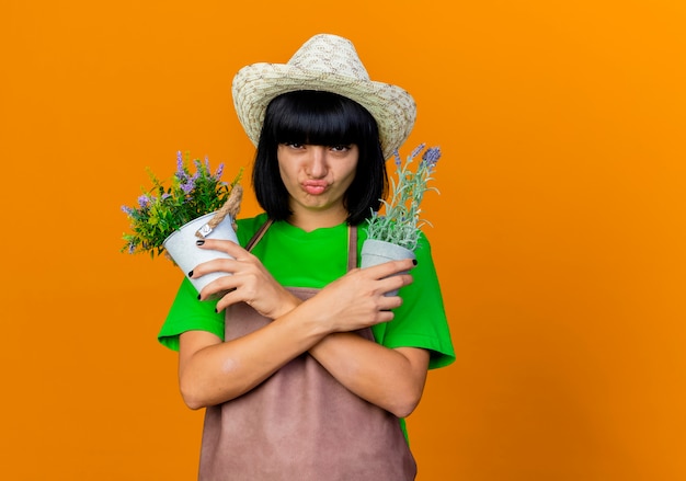 Il giovane giardiniere femminile scontento in uniforme che indossa il cappello da giardinaggio tiene i vasi da fiori