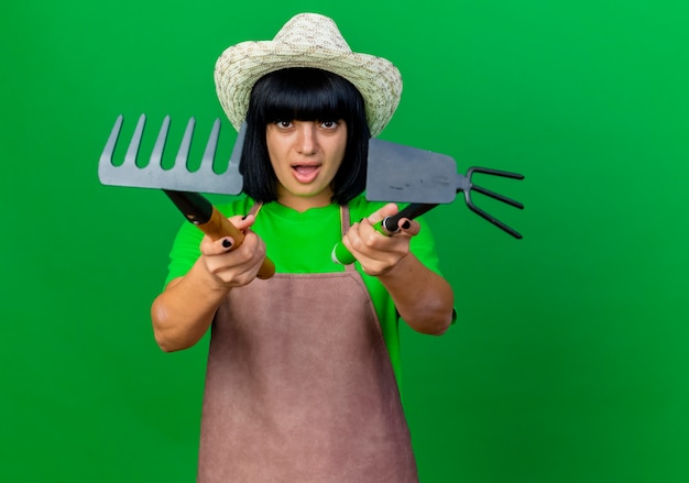 Foto gratuita il giovane giardiniere femminile scontento in uniforme che indossa il cappello da giardinaggio tiene il rastrello e il rastrello della zappa