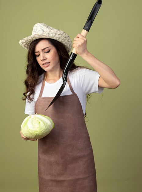 Unpleased young female gardener in uniform wearing gardening hat holds spade over cabbage isolated on olive green wall