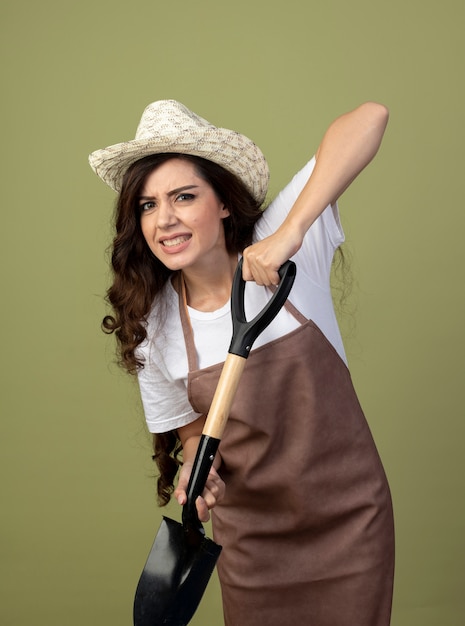 ガーデニング帽子をかぶって制服を着た不機嫌な若い女性の庭師は、オリーブグリーンの壁に隔離されたスペードを保持