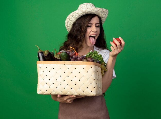 ガーデニング帽子を身に着けている制服を着た不機嫌な若い女性の庭師は、緑の壁に隔離された野菜のバスケットとトマトを保持している舌を突き出します