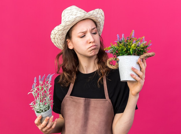 Una giovane giardiniera scontenta che indossa un cappello da giardinaggio che tiene e guarda i fiori in vasi da fiori isolati su una parete rosa