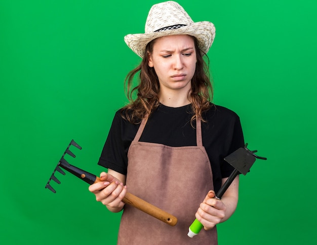 Unpleased young female gardener wearing gardening hat holding and looking at rake with hoe rake