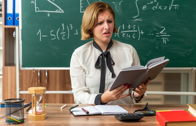 unpleased young female teacher sits at table with school supplies reading book in classroom