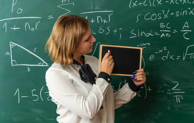 unpleased young female teacher standing in front blackboard holding and looking at mini blackboard with stranded for board in classroom