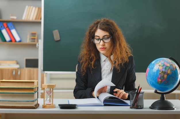 unpleased young female teacher wearing glasses flipping book sitting at desk with school tools in classroom