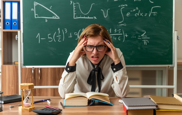 Foto gratuita una giovane insegnante scontenta con gli occhiali si siede al tavolo con gli strumenti della scuola mettendo le mani sulle tempie in classe