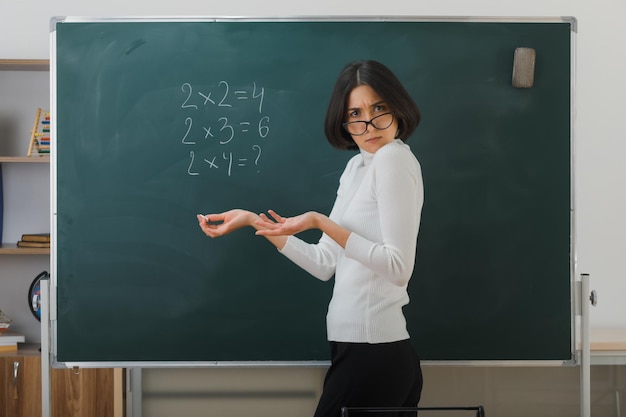 Free photo unpleased young female teacher wearing glasses standing in front blackboard and writes in classroom