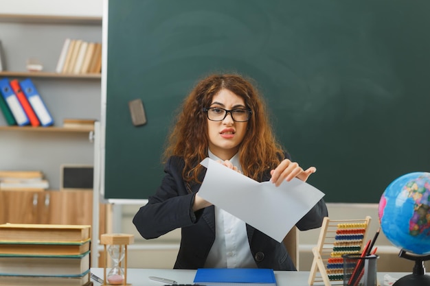 недовольная молодая учительница в очках рвет бумагу, сидя за партой со школьными инструментами в классе