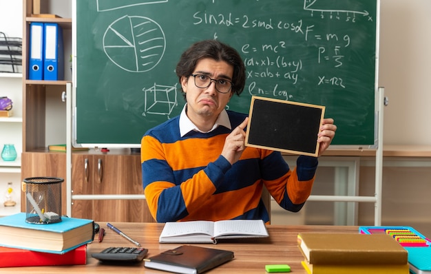 unpleased young geometry teacher wearing glasses sitting at desk with school supplies in classroom showing mini blackboard looking at front