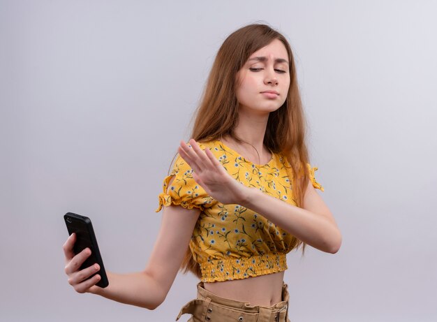 Unpleased young girl holding mobile phone and pointing with hand at it on isolated white wall