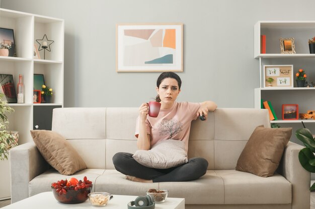 Unpleased young girl holding tv remote with cup of tea sitting on sofa behind coffee table in living room