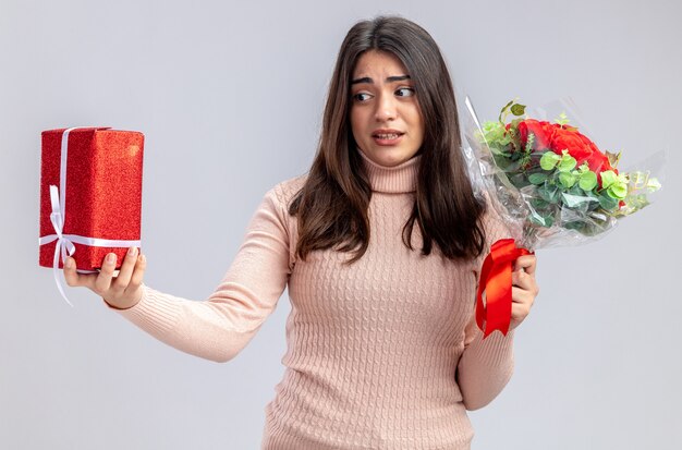 Unpleased young girl on valentines day holding bouquet looking at gift box in her hand isolated on white background