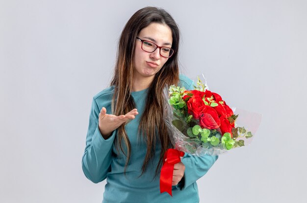 Unpleased young girl on valentines day holding and points with hand at bouquet isolated on white background