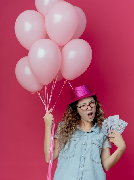 Недовольная молодая девушка в очках и розовой шляпе держит воздушные шары и деньги, изолированные на розовом