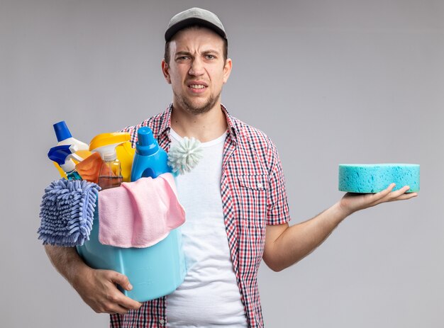 Unpleased young guy cleaner wearing cap holding bucket with cleaning tools and cleaning sponge isolated on white background