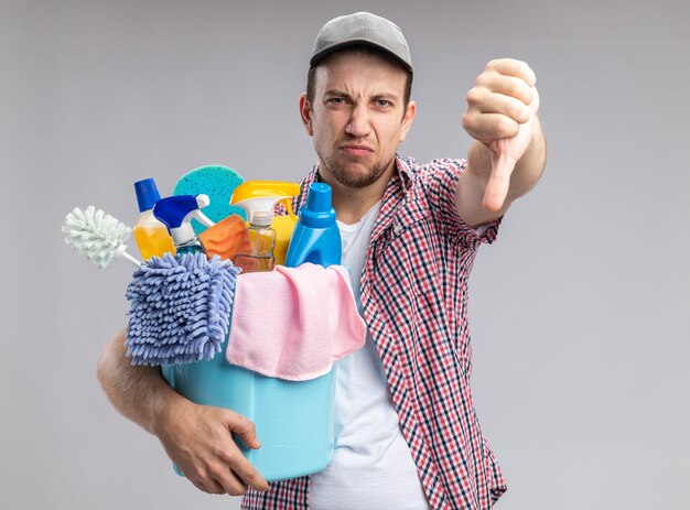 Недовольный молодой парень-уборщик в кепке держит ведро с чистящими инструментами, показывая большой палец вниз на белой стене