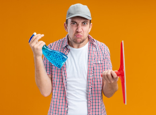 Free photo unpleased young guy cleaner wearing cap holding cleaning agent with mop head isolated on orange background