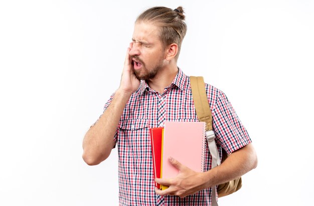 Unpleased young guy student wearing backpack holding books putting hand on aching tooth isolated on white wall