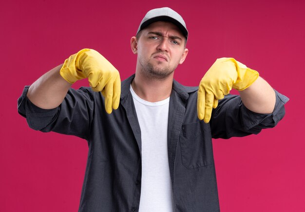 Unpleased young hamdsome cleaning guy wearing t-shirt and cap with gloves points at down isolated on pink wall
