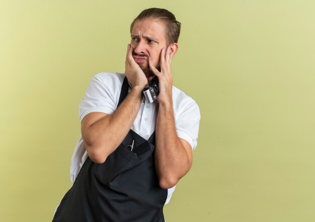 Unpleased young handsome barber holding hair clippers looking at side putting hands on face isolated on olive green background with copy space