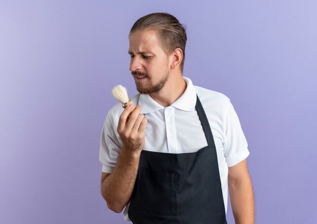 Unpleased young handsome barber wearing uniform holding and looking at shaving brush isolated on purple background with copy space