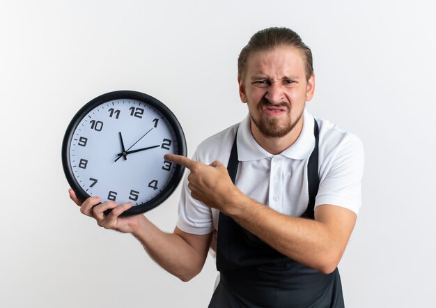 Unpleased young handsome barber wearing uniform holding and pointing at clock isolated on white background