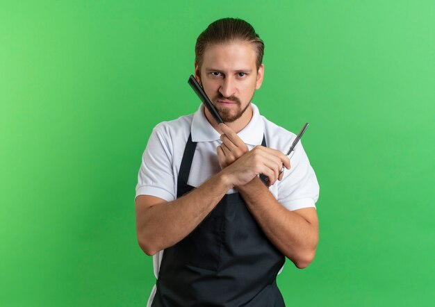 Unpleased young handsome barber wearing uniform holding scissors and touching his face with comb isolated on green background with copy space