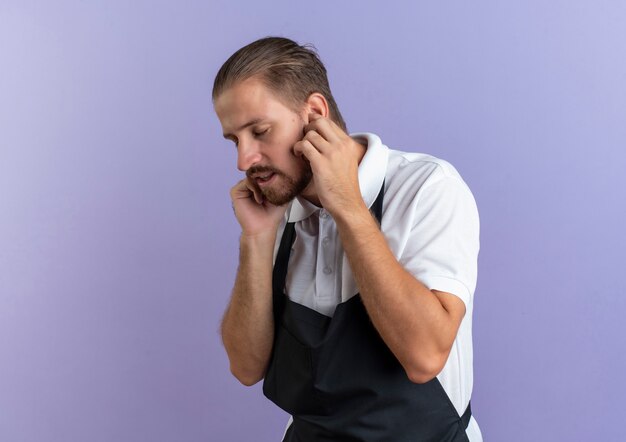 Недовольный молодой красивый парикмахер в униформе, засовывая пальцы в уши с закрытыми глазами, изолированными на фиолетовом фоне с копией пространства