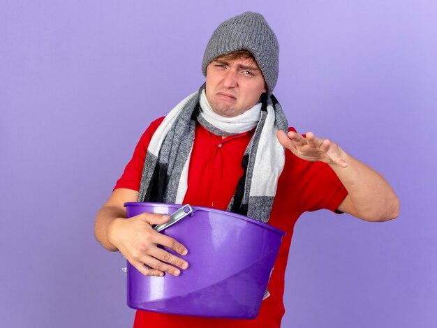 Недовольный молодой красивый блондин больной мужчина в зимней шапке и шарфе держит пластиковое ведро, держа руку в воздухе, изолированную на фиолетовой стене с копией пространства