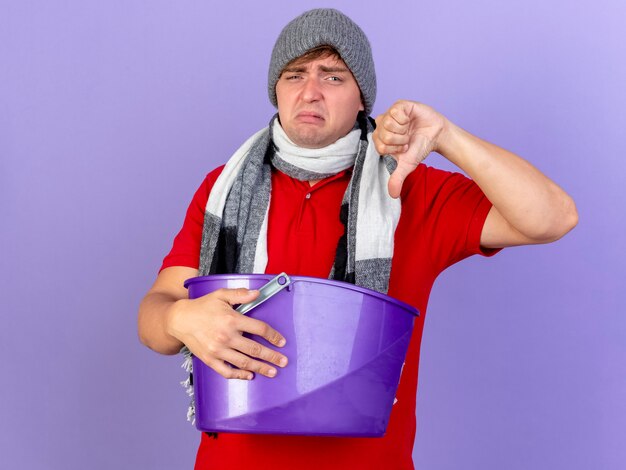 Unpleased young handsome blonde ill man wearing winter hat and scarf holding plastic bucket looking at camera showing thumb down isolated on purple background with copy space