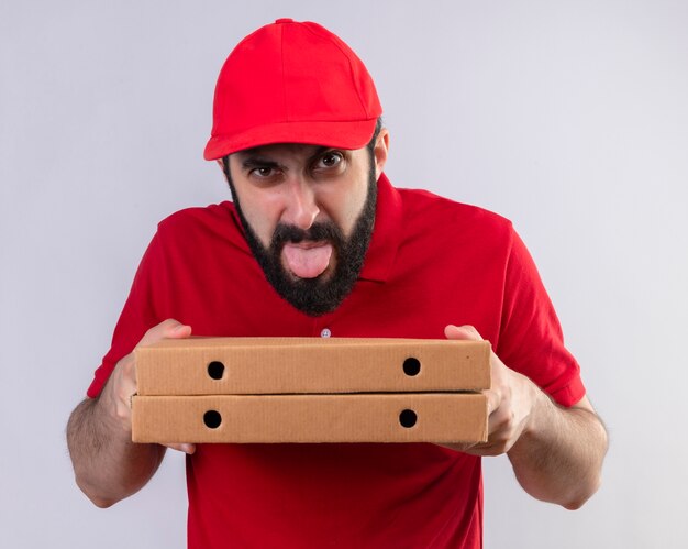 Unpleased young handsome caucasian delivery man wearing red uniform and cap holding pizza boxes and showing tongue isolated on white background