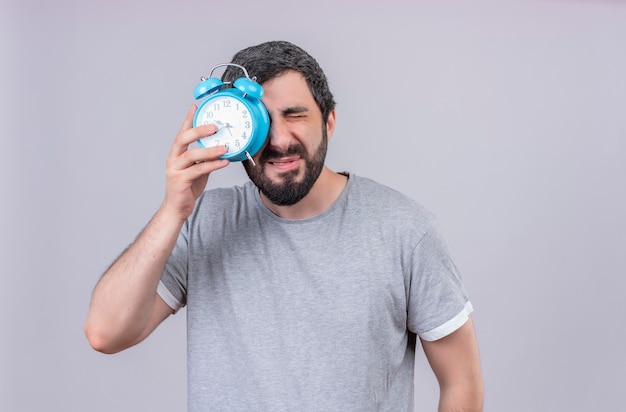 Unpleased young handsome caucasian man putting alarm clock on his eye and closing his eyes isolated on white background with copy space