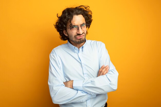 Unpleased young handsome caucasian man wearing glasses standing with closed posture looking at camera isolated on orange background with copy space