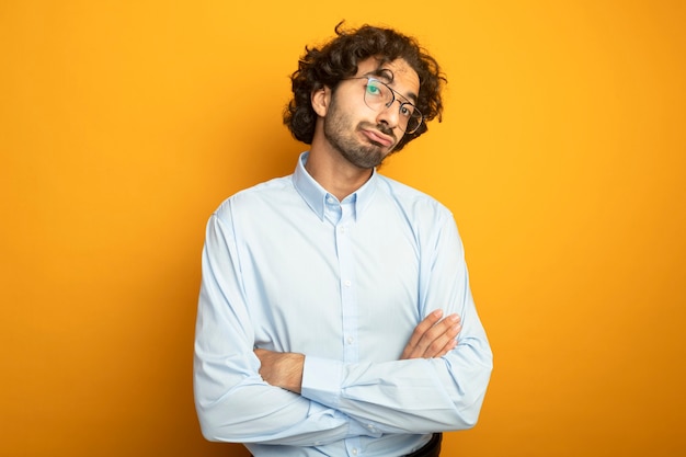 Unpleased young handsome caucasian man wearing glasses standing with closed posture looking at camera isolated on orange background with copy space