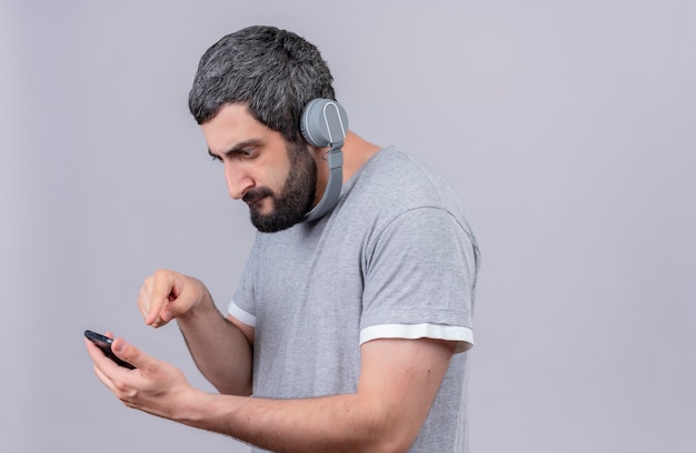 Giovane uomo caucasico bello dispiaciuto che indossa le cuffie guardando e utilizzando il suo telefono cellulare isolato su priorità bassa bianca con lo spazio della copia