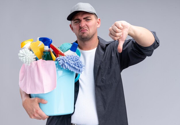 Недовольный молодой красивый уборщик в футболке и кепке, держащий ведро с инструментами для уборки, показывая большой палец вниз на белой стене