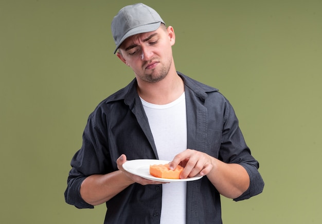 Free photo unpleased young handsome cleaning guy wearing t-shirt and cap holding plate with sponge isolated on olive green wall